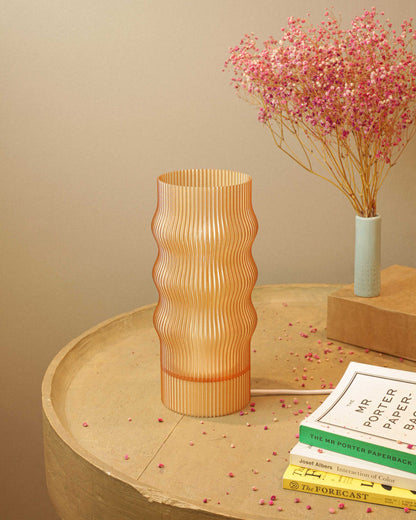 Brown table lamp on a round wooden table with books and a small plant in the background.