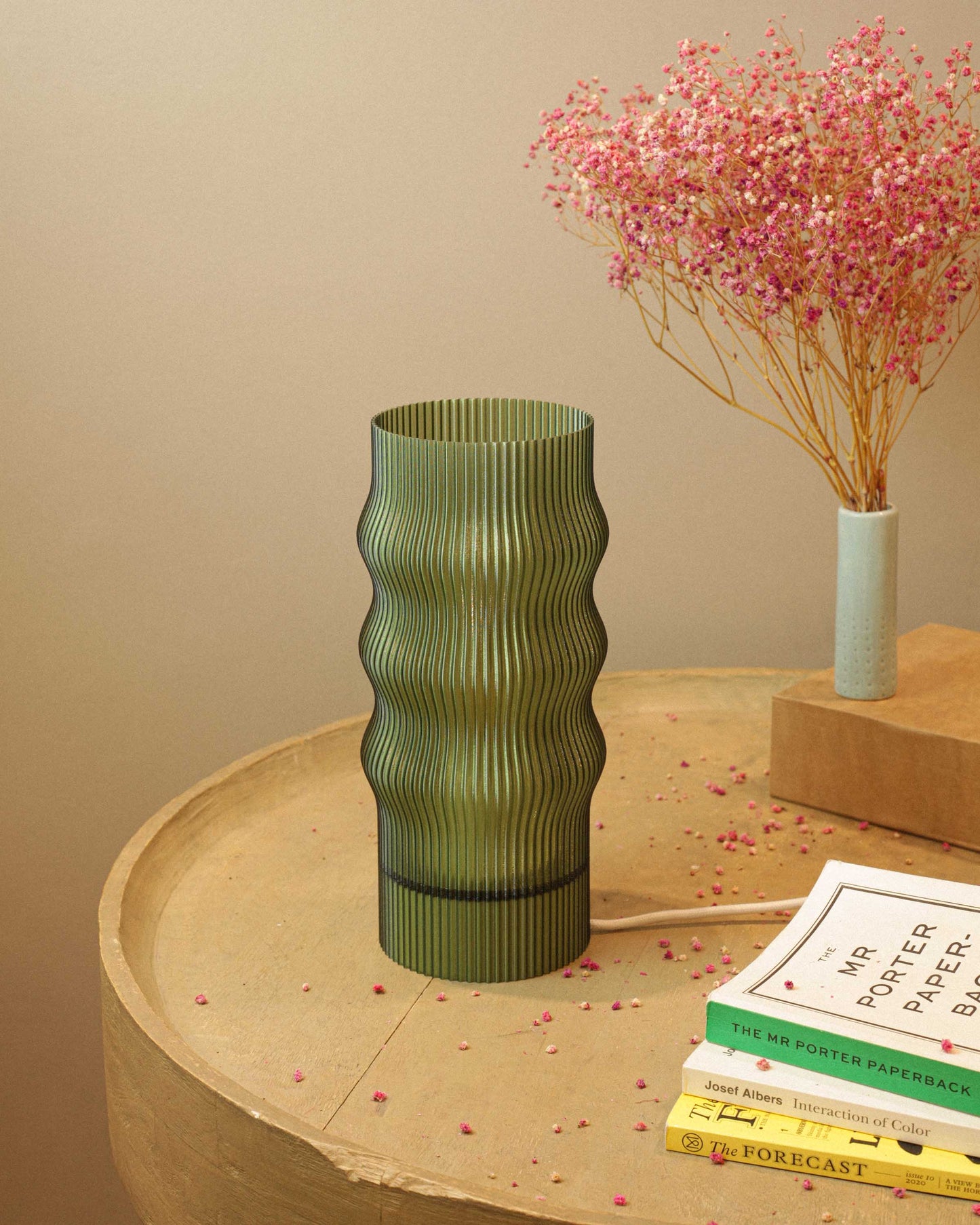 Green ribbed table lamp on a wooden surface with books and a small vase with pink flowers in the background.