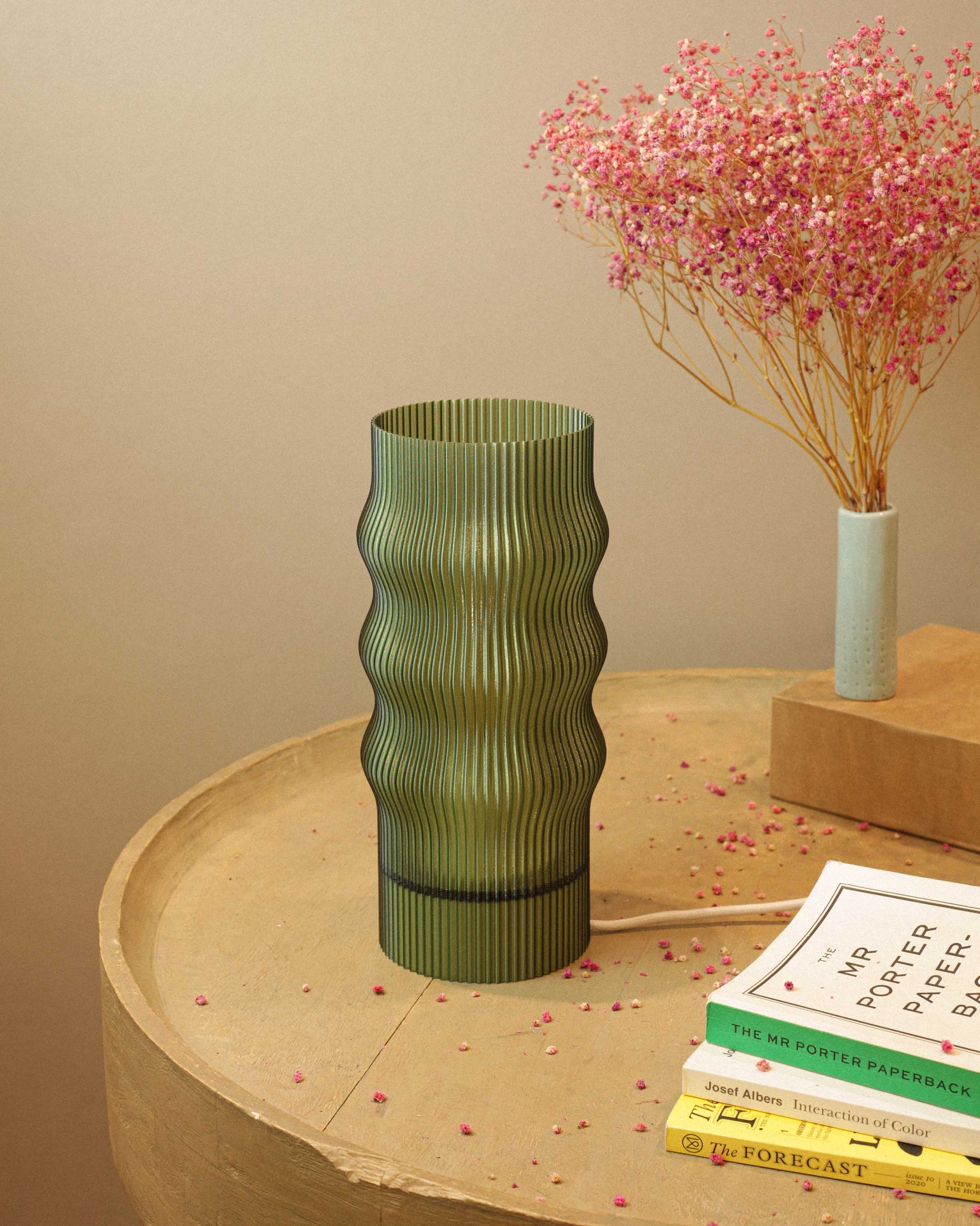 Green ribbed table lamp on a wooden surface with books and a small vase with pink flowers in the background.