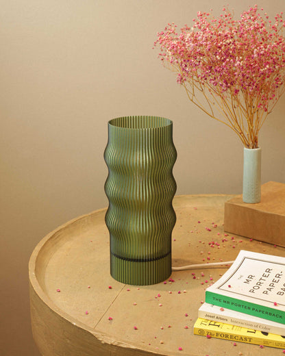 Green ribbed table lamp on a wooden surface with books and a small vase with pink flowers in the background.
